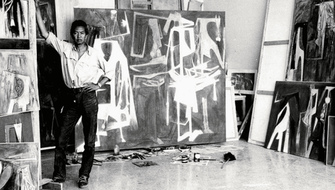 Artist Wilfredo Lam standing in his studio, gazing at the camera, surrounded by paintings leaning on