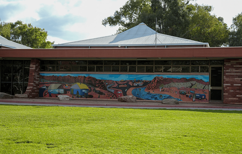 A one-story brick building on a green grass lawn decorated with a mural of a red desert rural landsc
