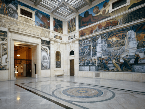 A photograph of murals decorating the walls of a marble museum lobby. The murals depict workers on a