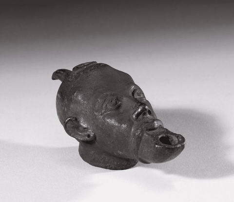 A blackened bronze oil lamp in the shape of a man's head, the open mouth is where the flame would co