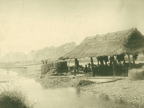 A group of workers sit under a thatched roof in a simple open-walled structure, perhaps resting on a
