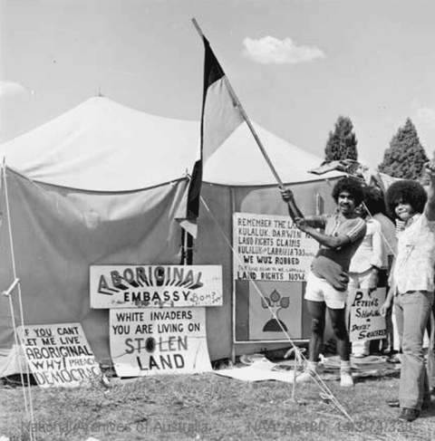 A black and white photograph of two activists raising a fist and a flag in front of a large light-co