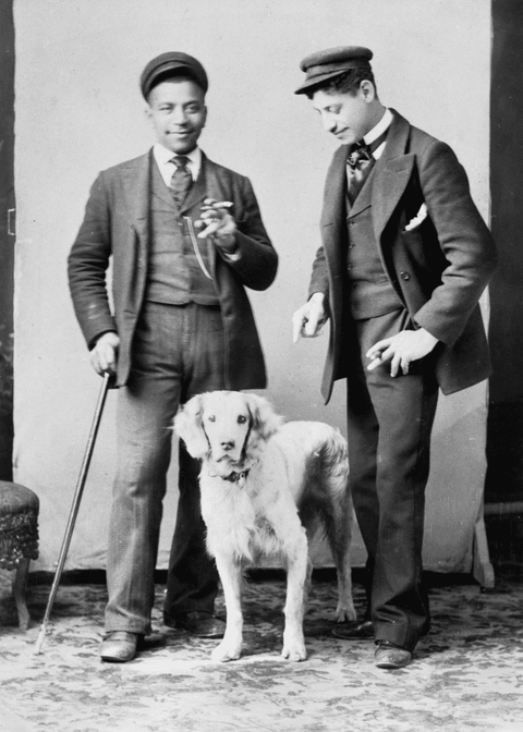 Full body portrait of a two unidentified African-American men wearing hats with a dog. Man on left h