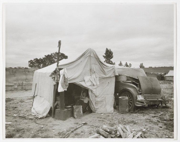 A black and white photograph depicts a temporary shelter made of sewn-together tarps draped over a d
